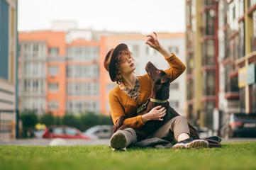Attractive stylish hipster girl in hat playing with dog sitting on lawn on blurred background with...