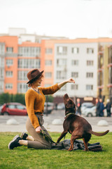 Stylish hipster girl dresses the dog on the lawn against the background of urban landscape, looks at the dog and smiles. Happy lady playing with dog on a walk. Vertical portrait of owner and dog