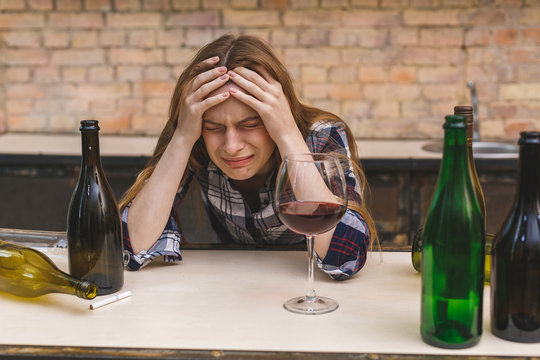 Young Sad And Wasted Alcoholic Woman Sitting At Kitchen Couch Drinking Red Wine, Completely Drunk Looking Depressed Lonely And Suffering Hangover In Alcoholism And Alcohol Abuse.