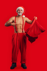 Christmas Freestyle. Young Santa Claus bare muscular upper body in hat standing isolated on red pointing inside gift bag smiling cheerful