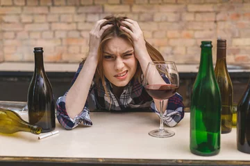 Fotobehang Young sad and wasted alcoholic woman sitting at kitchen couch drinking red wine, completely drunk looking depressed lonely and suffering hangover in alcoholism and alcohol abuse. © denis_vermenko
