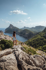 Epic shot of a woman hiking on the edge of a rock contemplating the incredible view of Cape Formentor in Majorca, Balearic Islands, Spain. Vertical photo.