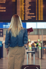 Young blond woman with suitcase and passport at airport against information board getting details about flight. - 298508051