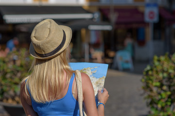 Fototapeta premium Young blonde beautiful girl in sun hat with map in her hands walking along European city, trying to find right way. Back view.