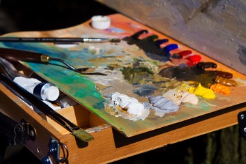 wooden artist palette with mixed fresh colorful oil paints in disorder, painter at work, outdoor...