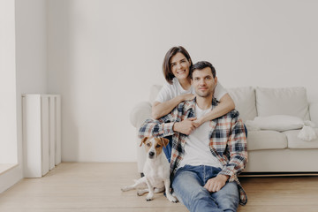 Portait of affectionate wife embraces husband who sits on floor near couch, their favourite pet poses at camera, make photo, being at home, buy new apartment, pose in living room with white walls