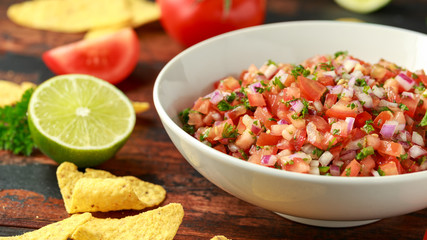 Mexican Tomato Salsa in white bowl with lime, red onion, jalapeno pepper, parsley and tortilla chips on wooden table