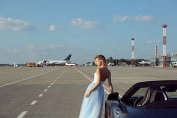 The elegant blonde beautiful woman posing near luxury vehicle on planes background. Girl wearing blue dress. No retouch.