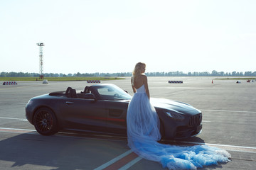 The elegant blonde beautiful woman posing near luxury vehicle on planes background. Girl wearing blue dress. No retouch.