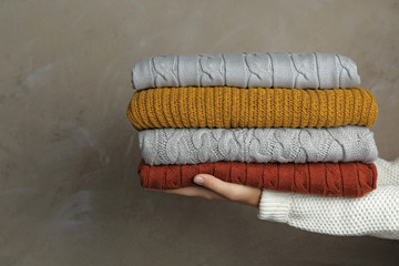 Woman holding stack of warm clothes on grey background, closeup. Autumn season