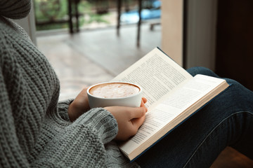 Woman with cup of coffee reading book near window indoors, closeup