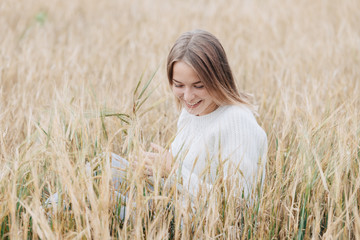 Beautiful young girl in a white sweater sits in a wheat field and smiles cheerfully.