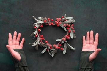 Female hands showing Heart-shaped decorative mistletoe Christmas wreath with red berries, flat lay...