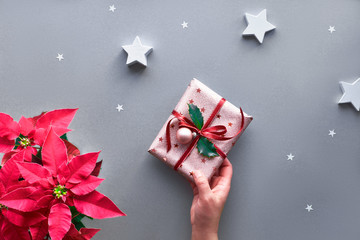 Female hand holding Christmas gift wrapped in metallic pink wrapping paper with red ribbon, holly leaf and bauble. Xmas creative flat lay on silver grey paper with red poinsettia and paper stars.
