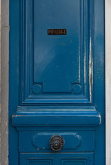 Old blue door. Bright blue color painted wooden door panel with round vintage doorknob and nameplate "Push" in French language. Details of geometric framed door with shabby surface. Door in Paris 