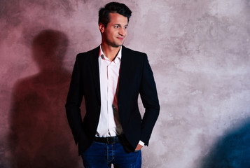 A man of her dreams. Attractive man with black hair and dark eyes is posing over the gray wall in a black jacket and jeans with his hands in the pockets, looking to the left and smiling slightly.