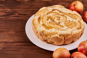 Baked apple pie on a white plate next to red apples on a brown wooden background