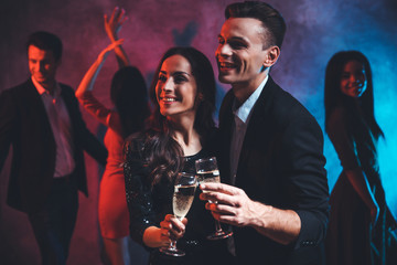 Vivid night. Alluring couple of a beautiful girl in a black shiny dress and an attractive man in a suit are posing with glasses of fizzy drink while their friends are dancing on the background.
