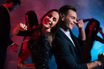 Party night. Beautiful girl with chestnut hair is dancing in a club back to back with handsome man in a suit, both are dancing with their hands forming fists.
