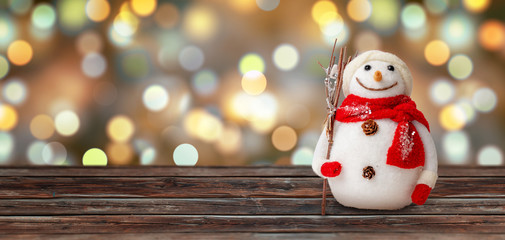 Snowman on wooden table. Christmas lights in background. Copy space beside for greeting text.