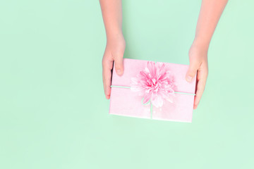 Hands holding a pink gift box with festive flower on top against pastel green background. Present and celebration or giveaway for feminine bloggers. Mothers Valentines Womens Day