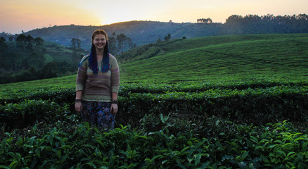 Naklejka na ściany i meble A white Caucasian young smiling woman with blue pigtails stands at sunset on a field with flowering bushes of tea. Tea farming and plantation in India.