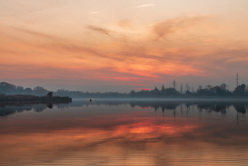 Obraz premium Beautiful sunset on the lake. A fisherman on a boat is fishing. Misty evening on the lake at sunset. Beautiful landscape with sunset and lake