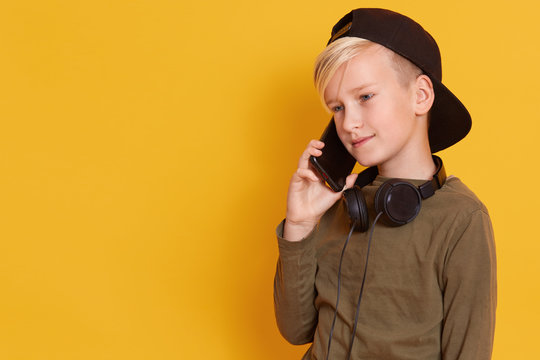 Close up portrait of blonde boy speaking by mobile phone and having pleasant facial expression, charming model posing isolated over yellow studio background. Copy space for advertisment or promotion.