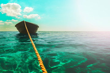 A thick clothesline is tied to an anchor that lies at the bottom of a clear blue sea, keeping the green boat on the surface in Sunny weather with clouds and glare, calm blue sea, small waves © Aliaksei