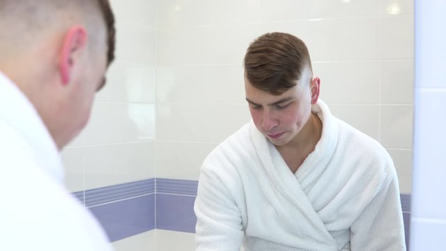A young man is brushing his teeth. A man in a white coat looked at his teeth in the mirror and started brushing them with a toothbrush. View through the mirror