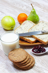 BREAKFAST WITH TOAST, CHOCOLATE COOKIES, JAM AND VARIED FRUIT WITH MILK GLASS ON LIGHT WOOD BACKGROUND