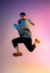 Fototapeta na wymiar Full length portrait of happy jumping man wearing casual clothes in neon light isolated on gradient background. Emotions, ad concept. Expressive hurrying up, late for work or sale, shopping.