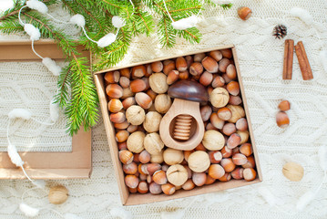 Obraz na płótnie Canvas Box with two kind of nuts in shell: walnuts, hazelnuts and mushroom shaped nutcracker on knitted background decorated with fir branch, cones and cinnamon sticks. Healthy Christmas gift concept