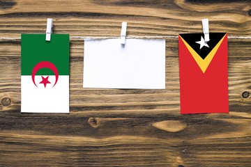 Hanging flags of Algeria and East Timor attached to rope with clothes pins with copy space on white note paper on wooden background.Diplomatic relations between countries.