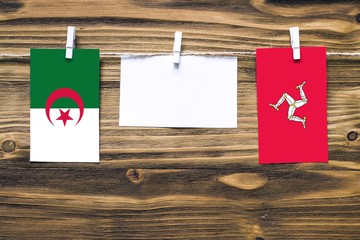 Hanging flags of Algeria and Isle Of Man attached to rope with clothes pins with copy space on white note paper on wooden background.Diplomatic relations between countries.