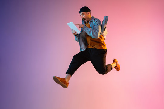 Full length portrait of happy jumping man wearing casual clothes in neon light isolated on gradient background. Emotions, ad concept. Using tablet, hurrying up, late for work or sale, shopping.