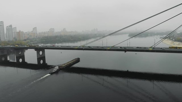 Big scow loaded with sand floating under concrete bridge through thick fog at industrial downtown. Barge sailing through industrialized smog near city, aerial drone view