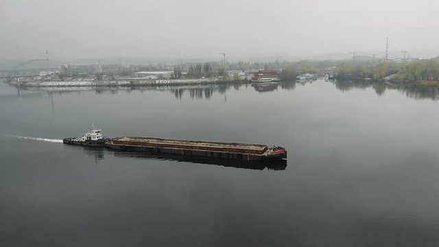 Big iron scow transporting sand through river in industrial downtown. Barge sailing on wide river through bridge in smog, aerial drone view