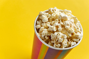 Popcorn in papercup. Multi coloured striped paper cup bucket with popcorn against a yellow background with copy space. Cinema, movies, streaming or entertainment concept. Dutch angle.
