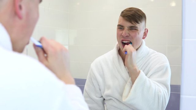 A young man is actively brushing his teeth. A man in a white bathrobe in front of a bathroom mirror. View through the mirror