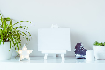 White scandinavian home interior design. Green plants in pots, star shape lamp, blank canvas on a stand, and stone detail on wall background. Minimalist simple hygge room decor. Close up. Copy space.