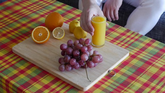 unrecognizable man with white pants sitting down and putting a glass of orange juice on table. this healthy meal is composed with Madras pattern tablecloth, wooden plate, raw fresh fruits.