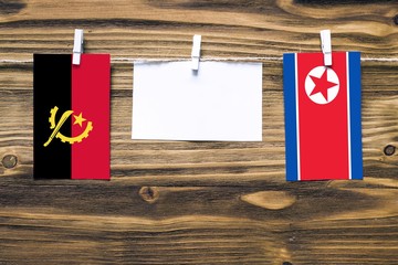 Hanging flags of Angola and North Korea attached to rope with clothes pins with copy space on white note paper on wooden background.Diplomatic relations between countries.