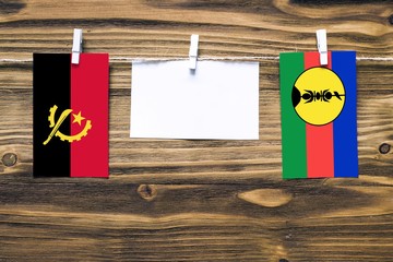 Hanging flags of Angola and New Caledonia attached to rope with clothes pins with copy space on white note paper on wooden background.Diplomatic relations between countries.