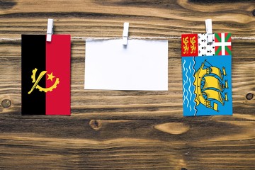 Hanging flags of Angola and Saint Pierre And Miquelon attached to rope with clothes pins with copy space on white note paper on wooden background.Diplomatic relations between countries.