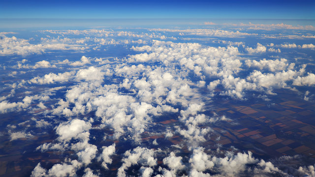 Flying over beautiful heaps of white clouds moving softly from right to left on the bright blue sky, and countryside landscapes far beneath. Picturesque view from the airplane window.