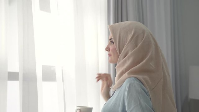 footage beautiful hijab woman enjoying coffee in the morning while opening the curtain to see the outdoors