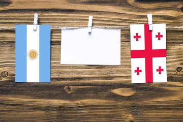 Hanging flags of Argentina and Georgia attached to rope with clothes pins with copy space on white note paper on wooden background.Diplomatic relations between countries.