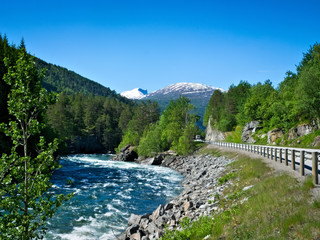 Fototapeta na wymiar A rushing mountain stream and a paved highway flow through rock and forest, with snow capped peaks and blue sky in the distance on a sunny summer day in Norway.