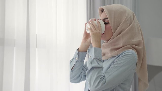 footage of beautiful Muslim women standing by the window feeling happy to enjoy coffee and looking the camera